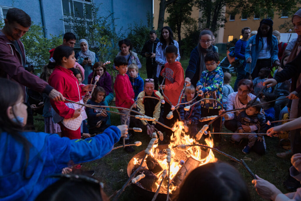 Grupo de crianças e adultos assa marshmallows em volta de uma fogueira durante evento ao ar livre à noite.